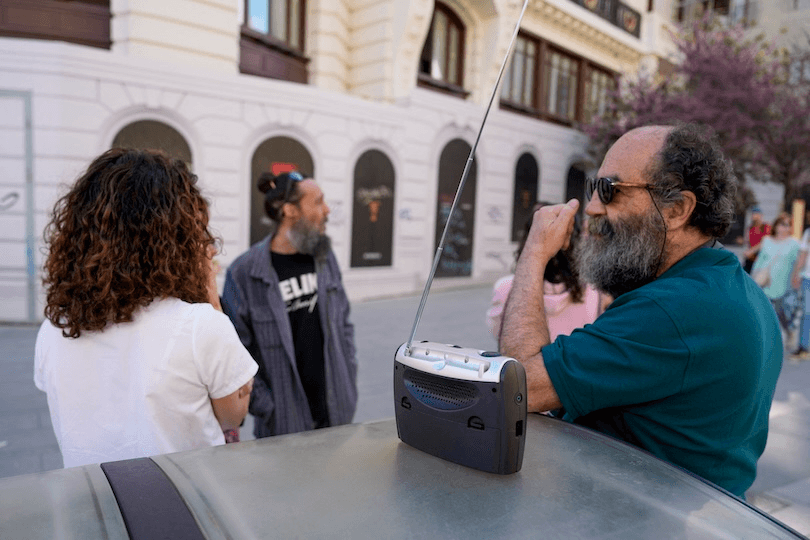 A man with sunglasses leans on a car with a portable radio, talking to two people on a city street. Buildings and trees are in the background.
