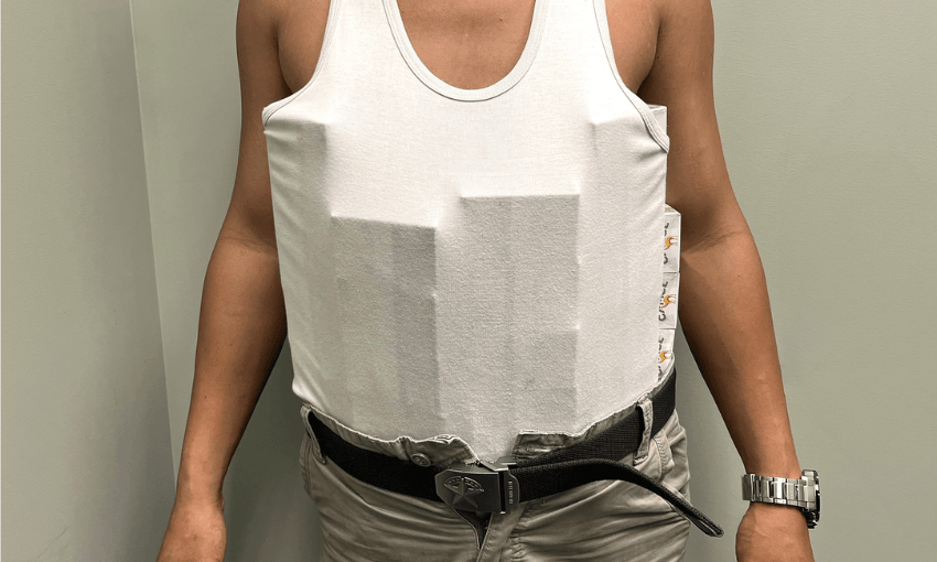 A person wearing a white tank top with two rectangular shapes visible underneath, which are cartons of cigarettes, along with beige pants, a black belt, and a silver wristwatch. The background is a plain light-colored wall.