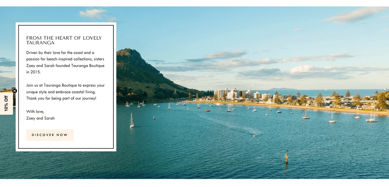 A scenic coastal view of Tauranga with calm blue waters, sailboats, a green mountain in the background, and waterfront buildings; an overlay text box shares the story of Tauranga Boutique’s founders, Zoey and Sarah.