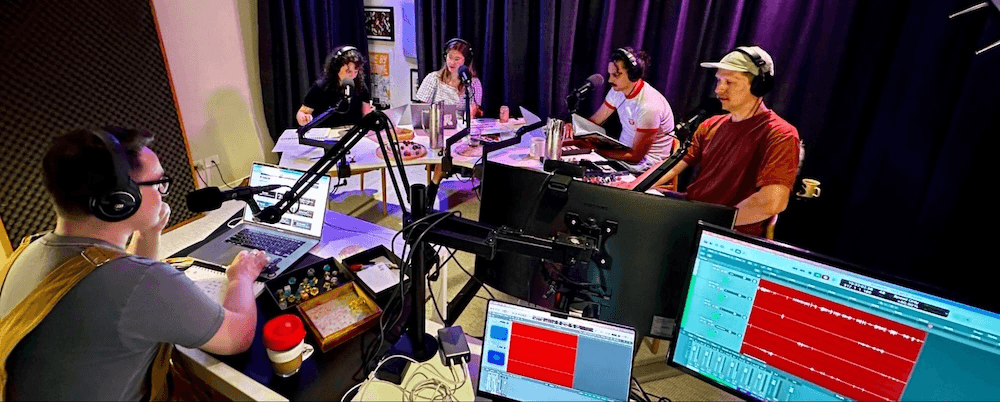 five people sit around a large table with headphones on, speaking into microphones