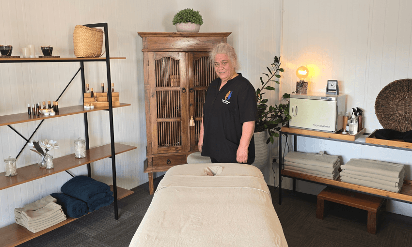 A massage therapist stands beside a massage table in a cosy, well-organised room with shelves holding towels and oils, a wooden cabinet, and soft lighting.