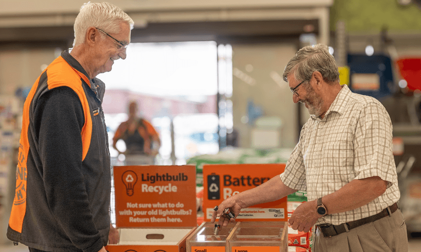a photo of a whitehaired man in a mitre 10 uniform observing a shorter grey haired man putting batteries into a battery recycling box