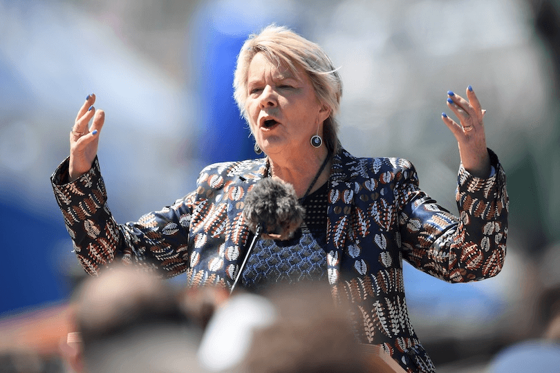 A woman with short blonde hair speaks passionately at an outdoor event, raising her hands. She wears a patterned jacket and stands behind a microphone, with blurred people in the foreground.