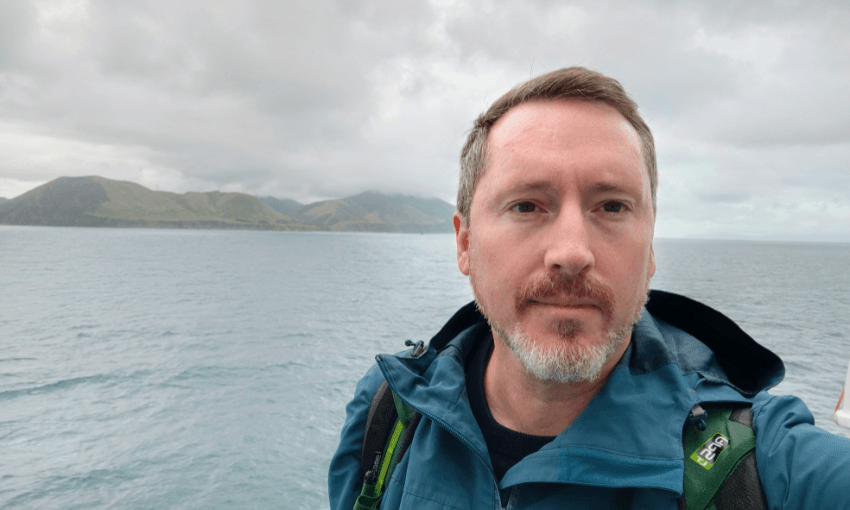 a white man with dark eyes and a white trimmed beard takes a selfie with the ocean and some hills behind him. 