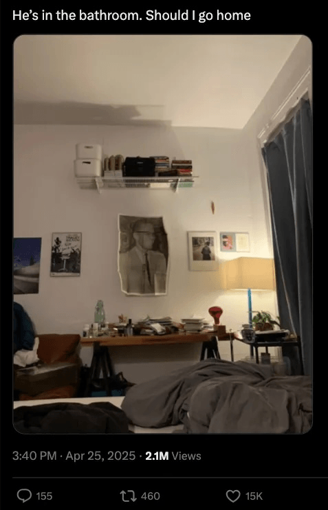 A dimly lit, cluttered bedroom with an unmade bed in the foreground, a messy desk with a lamp, books and art on the wall, and a tweet caption reading, “He’s in the bathroom. Should I go home”.