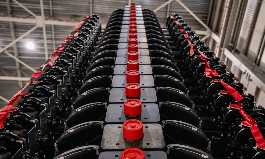 An image in a warehouse shows a stack of satellites all stacked up ready to be loaded into a rocket. The stacked up satellites fit together nicely and compactly, are black with red knobs on them.