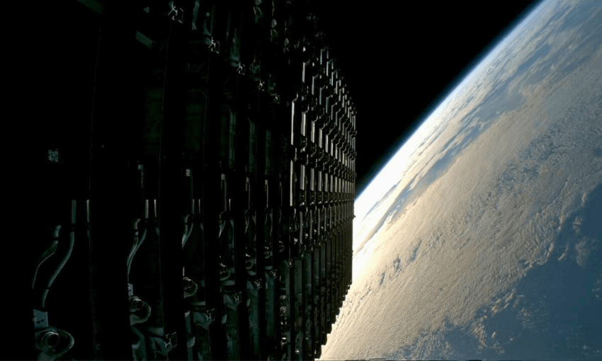 A stack of satellites or hardware panels in space, partially illuminated, with the curved horizon of Earth and a layer of clouds visible against the darkness of space in the background.
