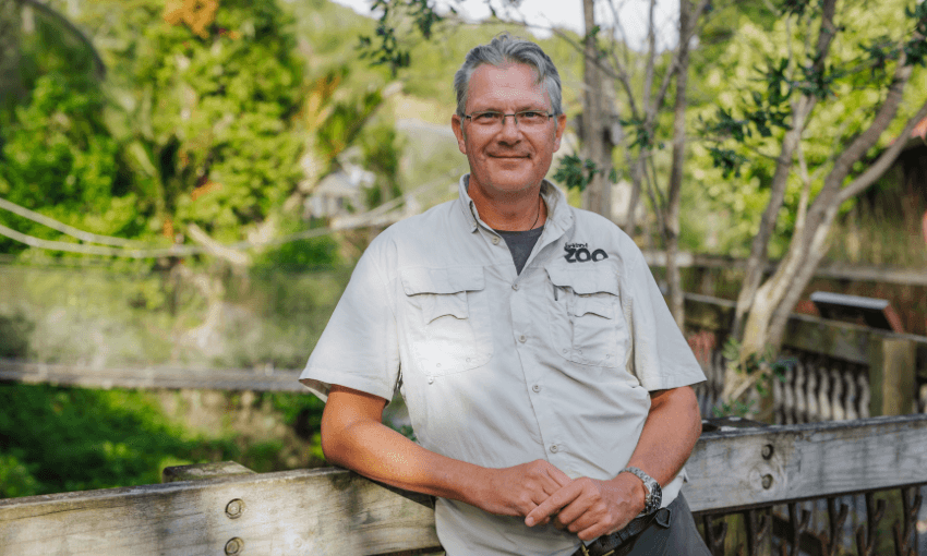 A man wearing glasses and a light khaki zoo shirt stands and smiles, resting his arm on a wooden fence with lush greenery and trees in the background.