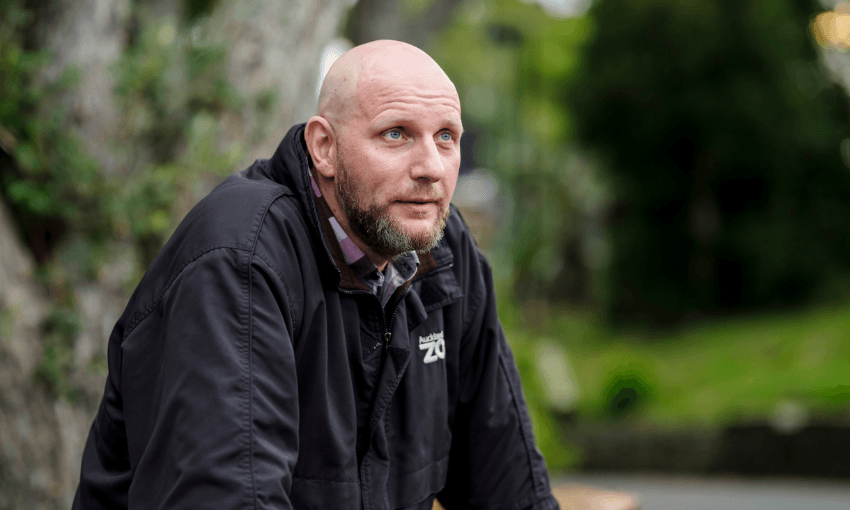 A bald man with a beard wearing a black jacket leans forward outdoors, looking thoughtfully into the distance. Trees and greenery are blurred in the background.