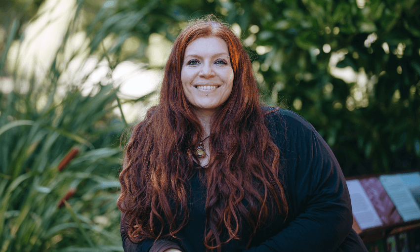 A woman with long red hair and a black top smiles while sitting outdoors, surrounded by green plants.