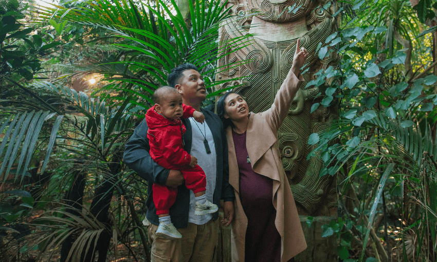 A family of three stands in a lush, green forest near a carved wooden pole. The father holds a child dressed in red, while the mother points upward, drawing attention to something among the leaves.