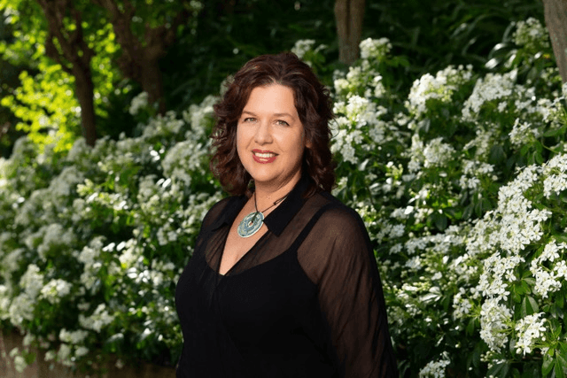 a white woman in a black top with a floral bush behind her