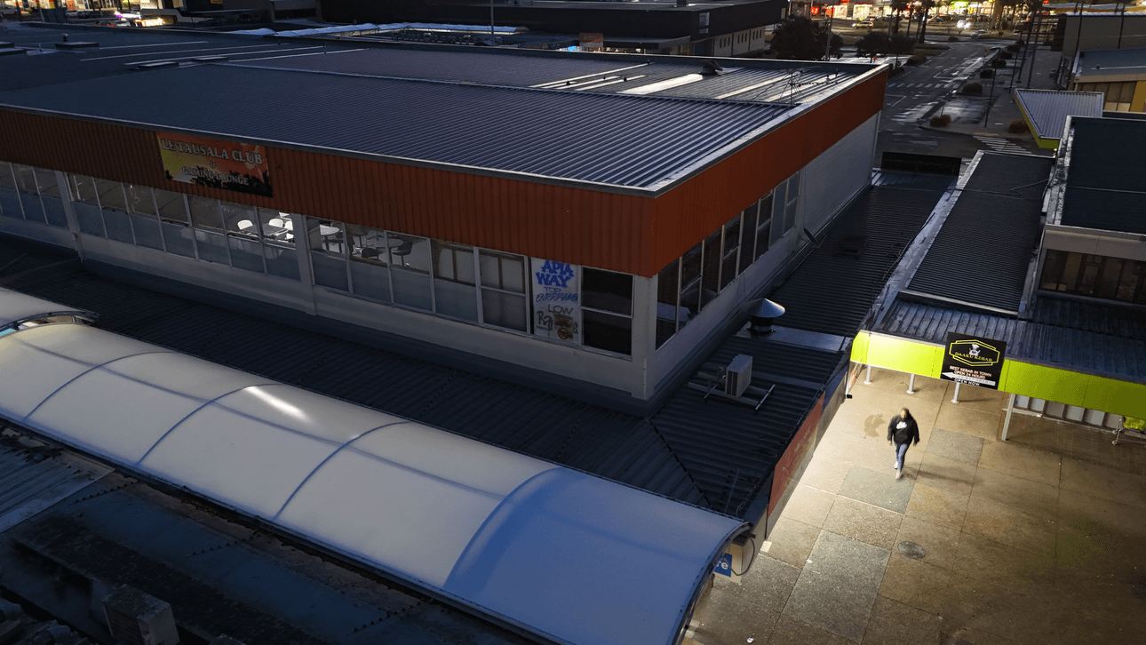 A person walks alone under a lit awning at night beside a large, modern commercial building with orange siding and many windows. The surrounding area appears quiet and mostly empty.