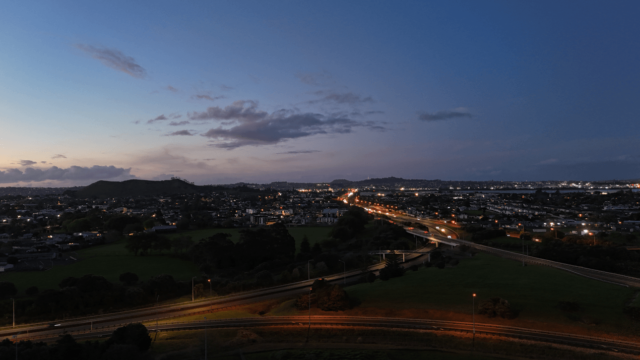 A cityscape at dusk with a winding road lit by car headlights, green fields in the foreground, and scattered houses. The sky is transitioning from blue to night with clouds and distant hills on the horizon.