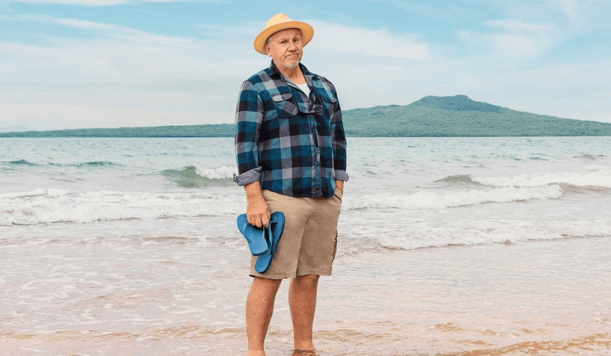 An older man stands barefoot on a beach, holding blue sandals. He wears a straw hat, plaid shirt, and khaki shorts. Gentle waves and a green, hilly island are visible in the background under a partly cloudy sky.