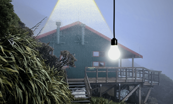 A green backcountry hut set against the side of a mountain on a foggy night. An electric lightbulb hangs down from the top of the frame