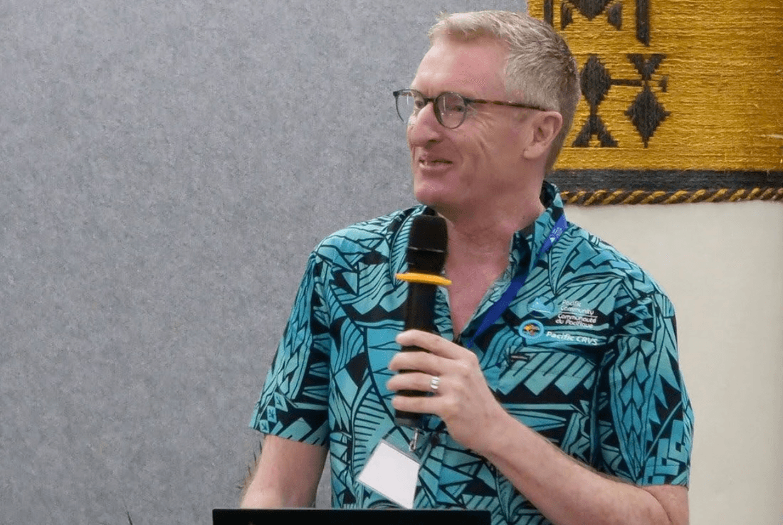 A smiling man in glasses and a blue shirt with Pacific patterns speaks into a microphone at an event, standing in front of a gray wall and a decorative yellow tapestry.