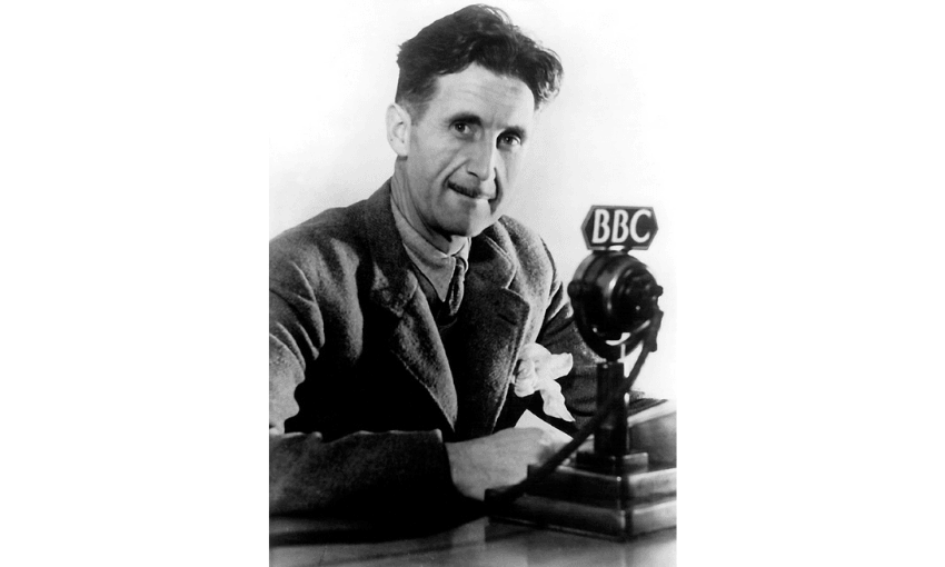 A black and white photograph of George Orwell in 1940 - he is in front of a microphone with a BBC label on it, and looking at the camera and looks middle aged.