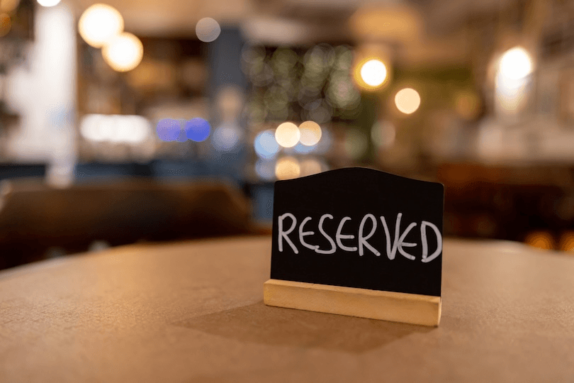 A small black sign with the word "RESERVED" written in white chalk sits on a round table in a softly lit, blurred restaurant or café setting.