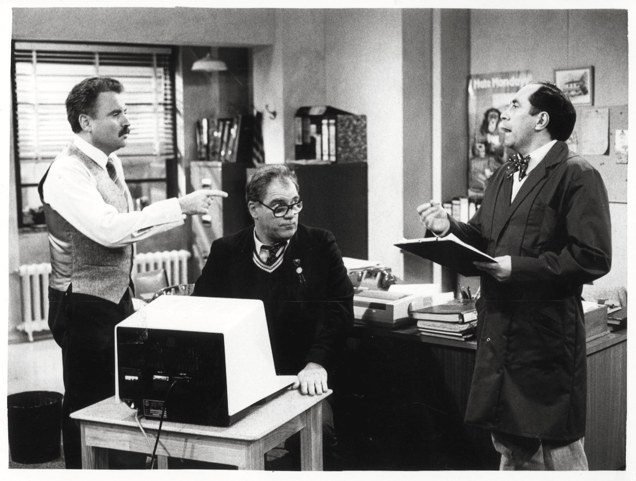 Three men in an office: one standing and pointing, one sitting at a desk with a computer, and another standing with a clipboard, engaged in conversation. The room has shelves, books, and office equipment.