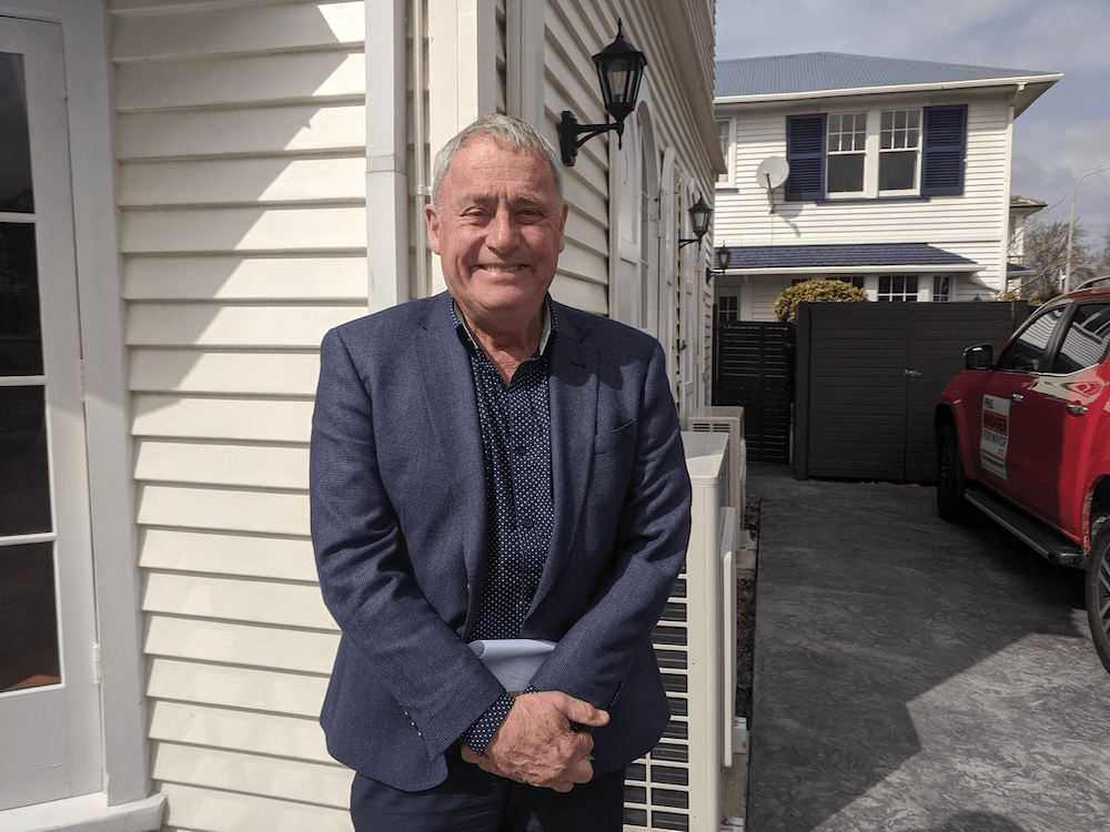 an image of an older white man in a blue suit smiling while holding a folder in front of a weatherboard house