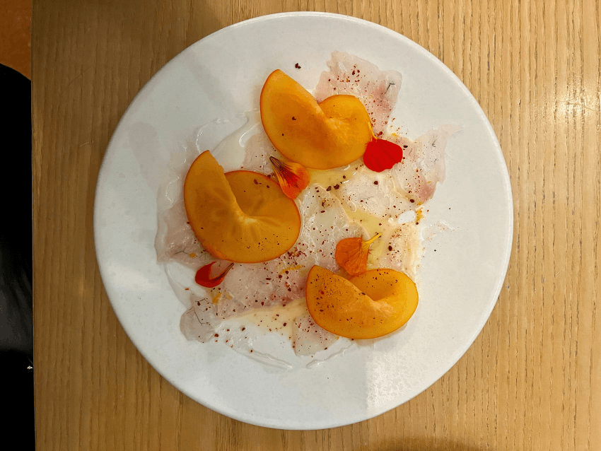 A white plate with thin slices of raw fish topped with orange persimmon slices, edible flower petals, and seasoning, placed on a light wooden table.