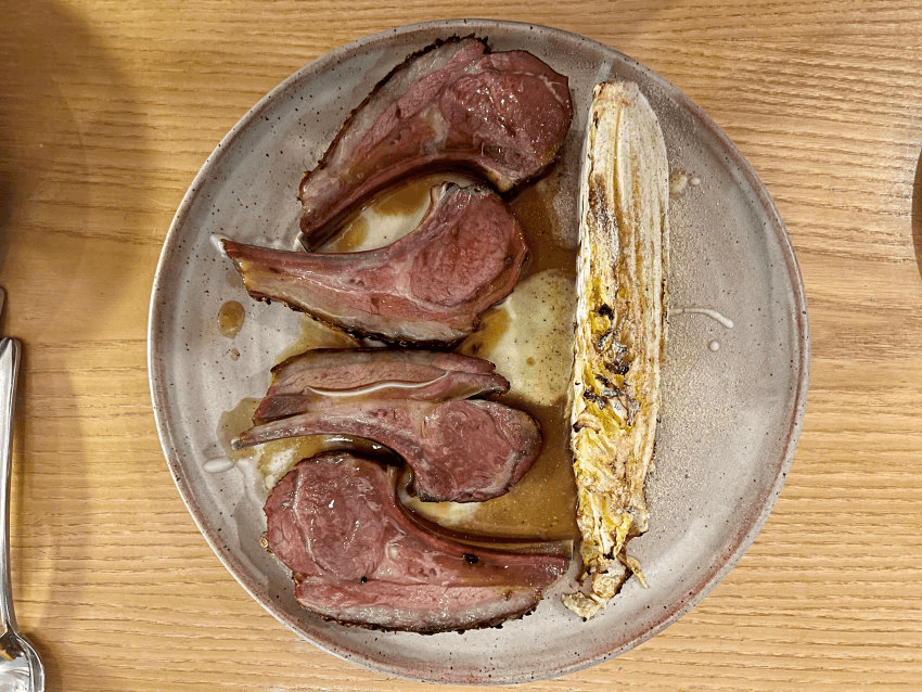 A plate with four cooked lamb chops arranged in a semicircle and a piece of grilled endive, all served on a light brown ceramic plate on a wooden table.