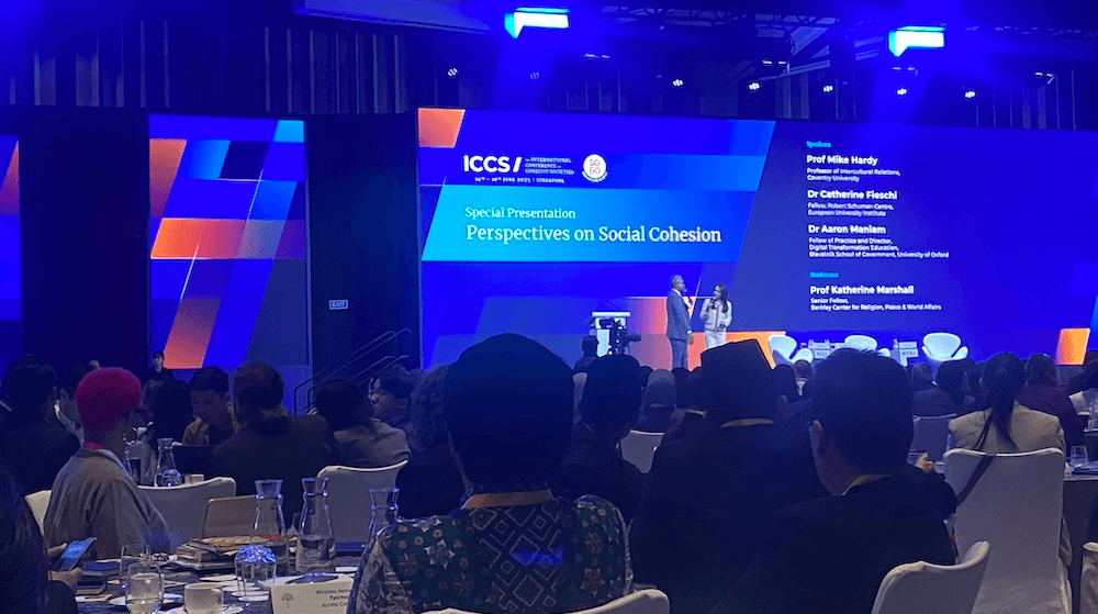 A faraway photo of a conference stage in a dimly lit function room, with a large blue screen at the back and two speakers on the stage