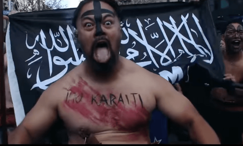 A man with face paint, body paint, and "IHU KARAITI" written on his chest sticks out his tongue while standing in front of a black flag with white Arabic script, with others similarly painted in the background.
