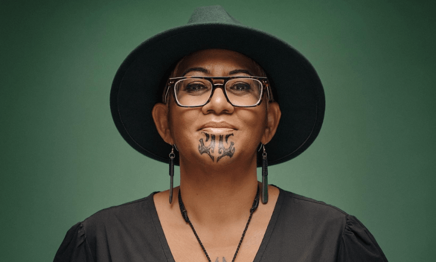 A person wearing a wide-brimmed dark hat, glasses, and earrings, with a traditional Māori chin tattoo and a confident expression, poses against a solid green background.