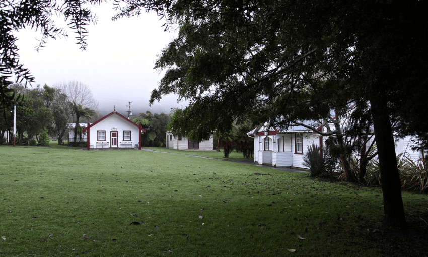 A grassy lawn with two traditional white wooden buildings, one with a red roof, surrounded by trees and shrubs on a cloudy day.