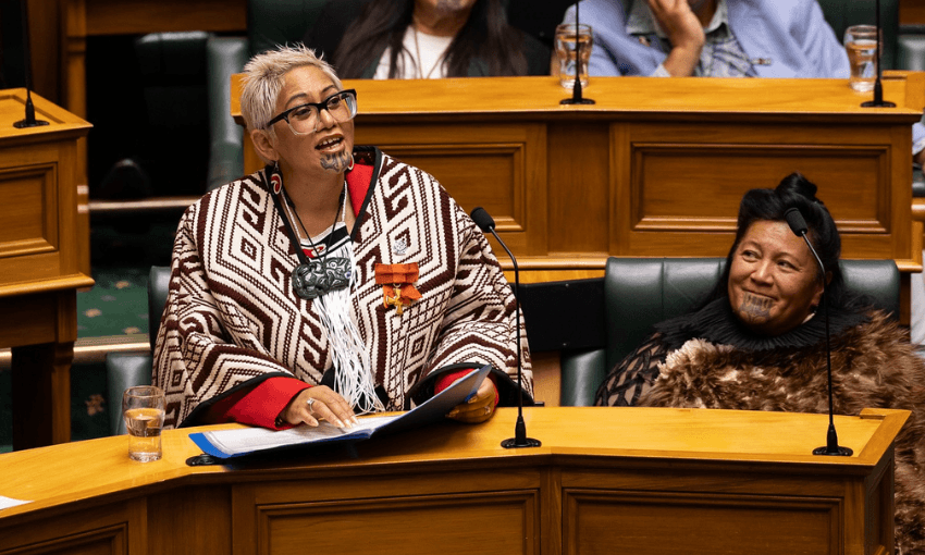 Two Māori women sit in a parliamentary chamber wearing traditional clothing and moko kauae (chin tattoos). One is speaking or smiling, holding papers, while the other looks on. Both appear engaged and dignified.