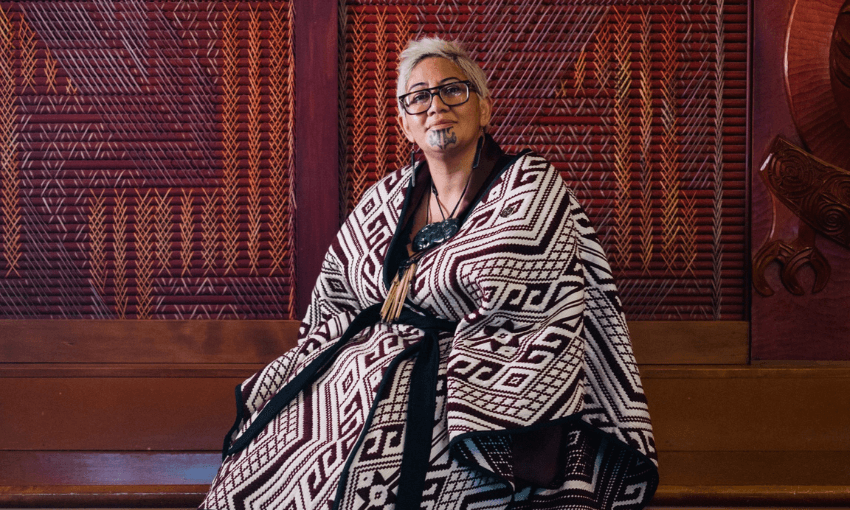 A person with glasses and traditional facial markings sits wrapped in a patterned cloak, in front of a wall with intricate wood carvings and patterns.