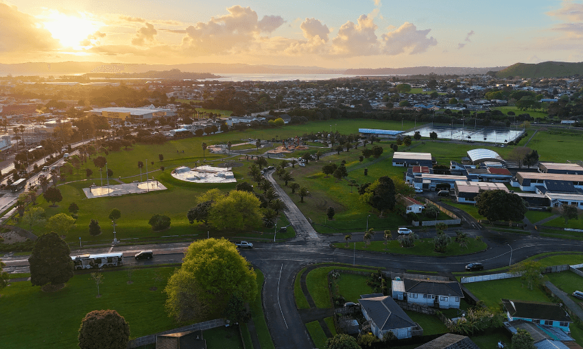 Aerial view of a suburban neighborhood at sunset, featuring green parks, sports fields, scattered houses, and buildings with trees and roads throughout the landscape. The sun is low on the horizon, casting a warm light.