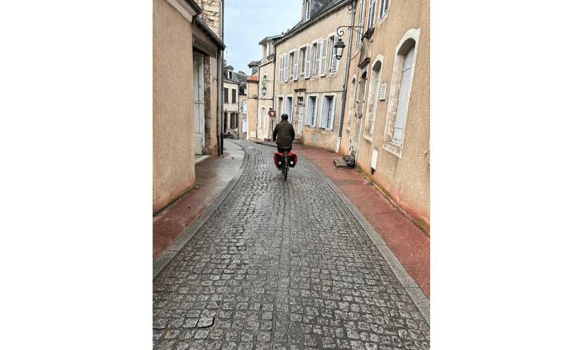 Photograph of person on a bike cycling down a cobbled street with joined houses on both sides, coloured peach.