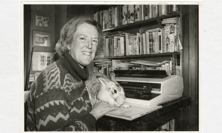 A black and white photo of the writer Margaret Mahy. She is sitting in a room with a lot of bookshelves. She's sitting at a desk with a typewriter in front of her, and a large rabbit on her knees. She is smiling.