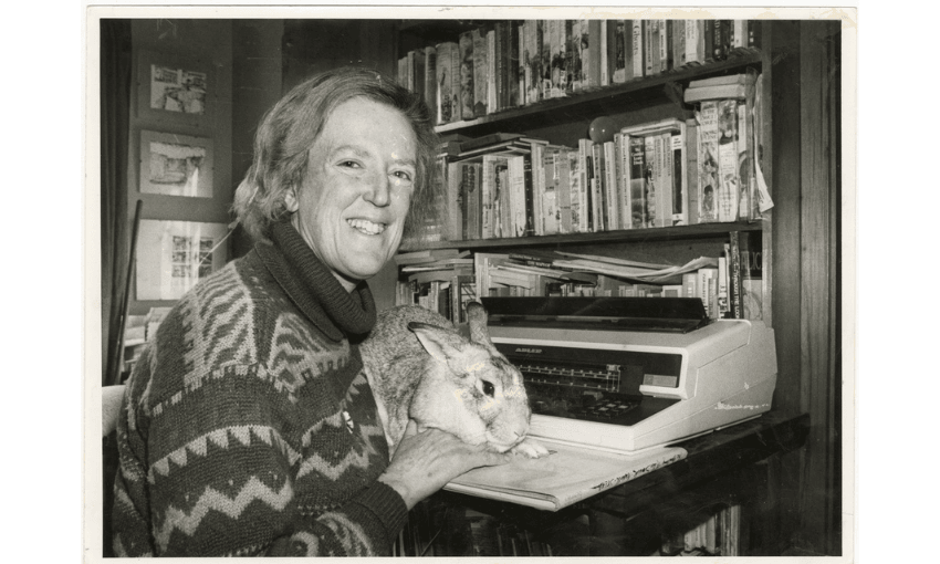 Black and white photo of Margaret Mahy sitting at her desk with a typewriter and a large rabbit on her knee.