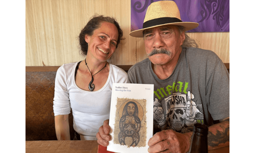 A man and a woman, both Māori, are seated next to each other. The man is holding up a book and they are both smiling at the camera.