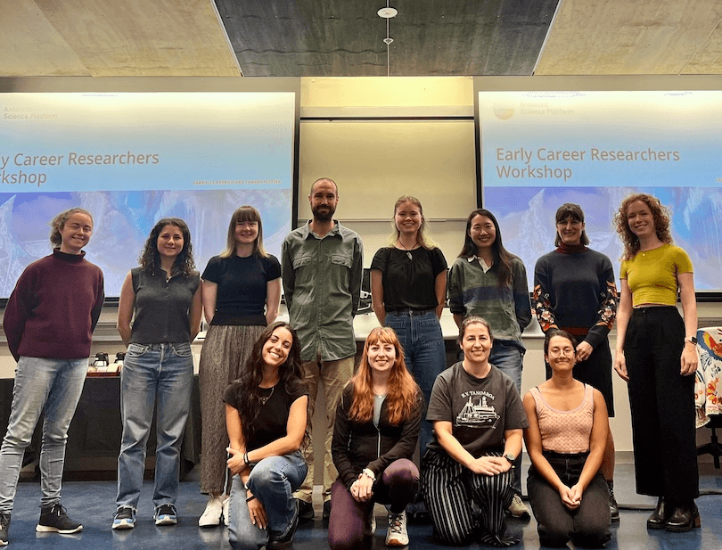 A group of 13 people pose and smile in a classroom in front of two screens that read "Early Career Researchers Workshop." The screens display blue and white graphics. Some people are standing, others are kneeling.