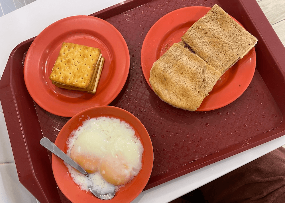 A red tray holds three red plates: one with two soft-boiled eggs, one with two slices of toasted bread, and one with a sandwich made of cream crackers. A metal spoon rests in the egg plate.
