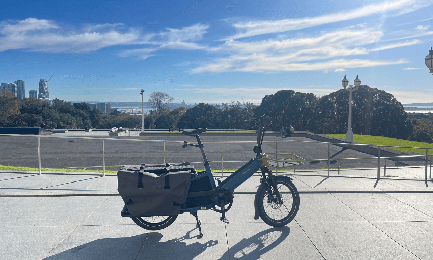 A cargo bicycle with panniers is parked on a paved area in the foreground, with trees, lamp posts, and a city skyline in the background under a blue sky with wispy clouds.