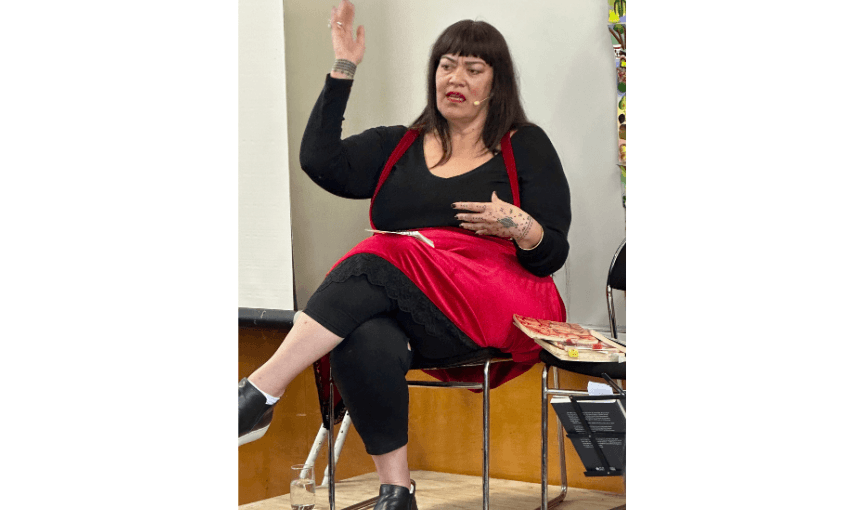 Photo of a Pasifika woman with her hand in the air, she has her mother open as she is speaking. She is seated and wearing black and red clothes. 