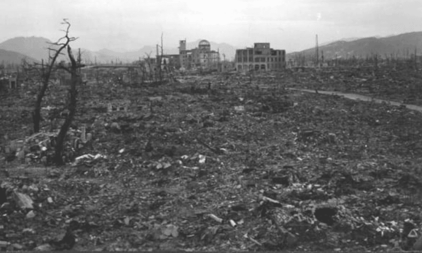 A black-and-white photo showing a devastated landscape with rubble, barren trees, and destroyed buildings in the distance, illustrating the aftermath of an atomic bomb explosion.