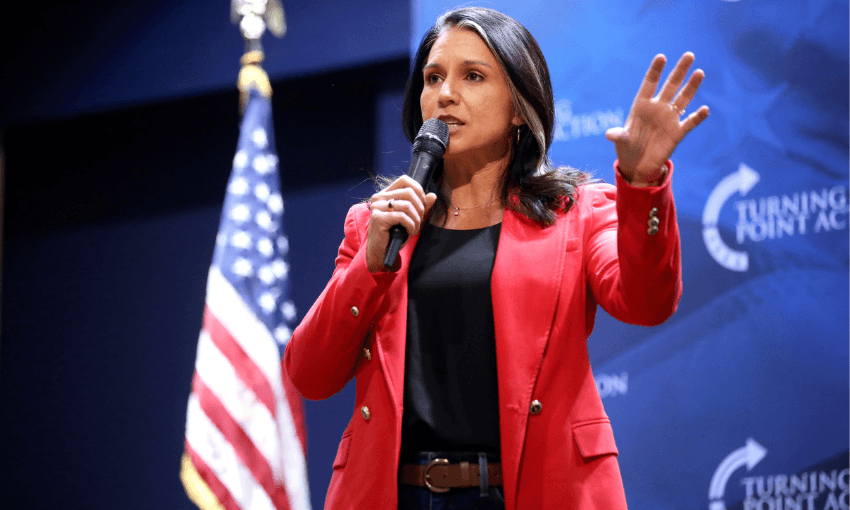 A woman (Tulsi Gabbard) in a red blazer speaks into a handheld microphone on stage, gesturing with her left hand. Behind her is an American flag and a blue backdrop with “Turning Point Action” logos.