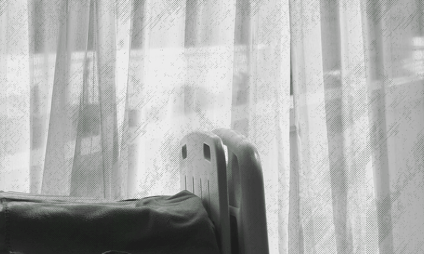 A black and white photo of a hospital bed partially visible in front of sheer curtains, with soft daylight coming through the window.