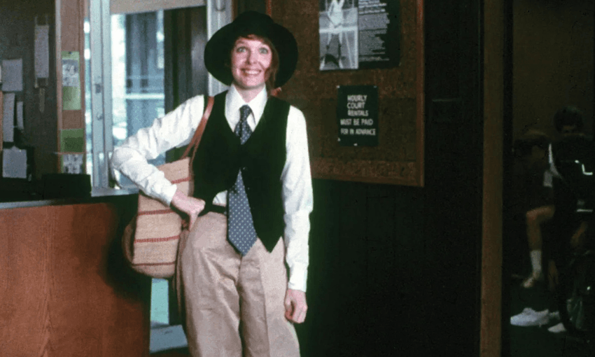 A person (Diane Keaton in Annie Hall) wearing a wide-brimmed hat, vest, tie, white shirt, and khaki pants stands indoors, smiling and holding a striped shoulder bag. There is a sign and doorway in the background.