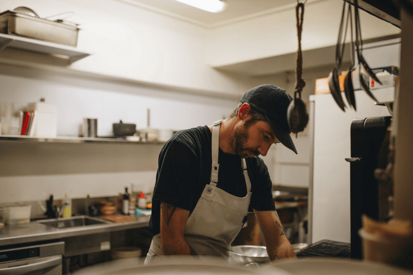 A person wearing a black shirt, white apron, and dark cap is working in a commercial kitchen, focused on preparing food. Stainless steel shelves and kitchen equipment are visible in the background.