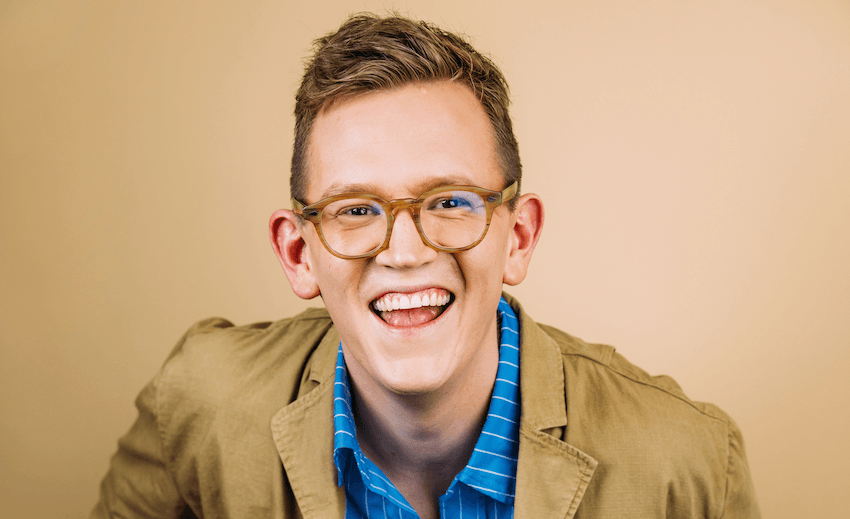 Chris Parker in glasses, a blue shirt and a beige jacket smiles in front of a beige background