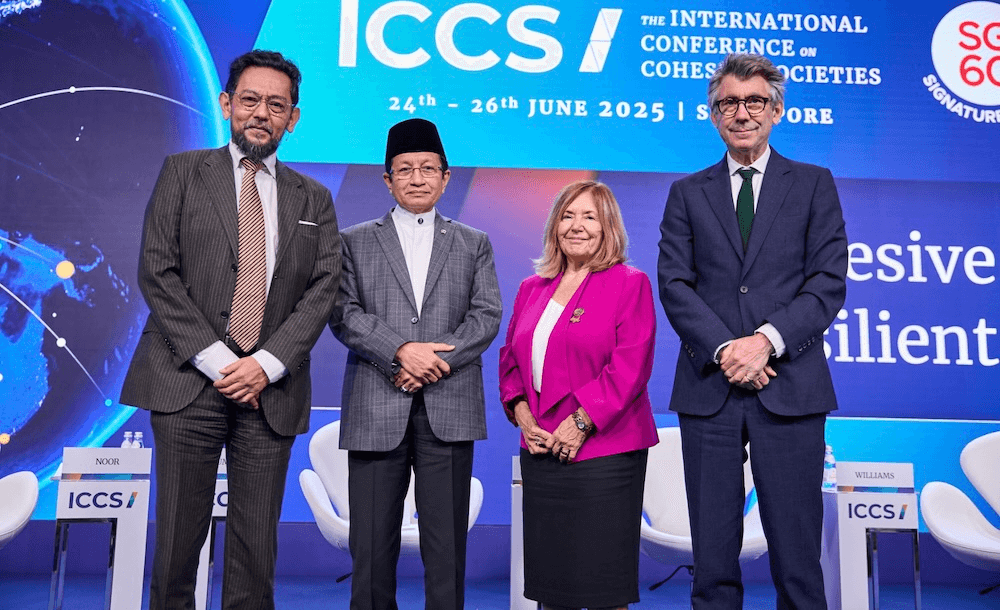 Four panelists in formal attire stand on stage at the International Conference on Cohesive Societies 2025, with a blue background displaying event details and empty chairs behind them.
