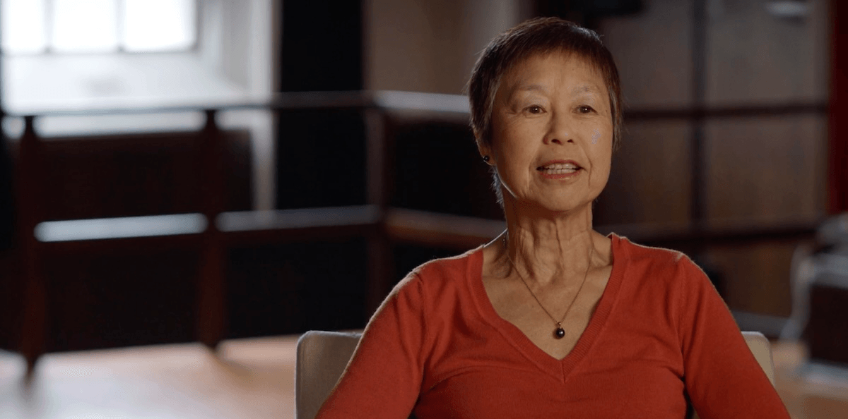 A woman with short brown hair and a red sweater sits indoors, speaking while looking slightly off-camera. The background is softly lit with windows and dark wooden furniture visible.